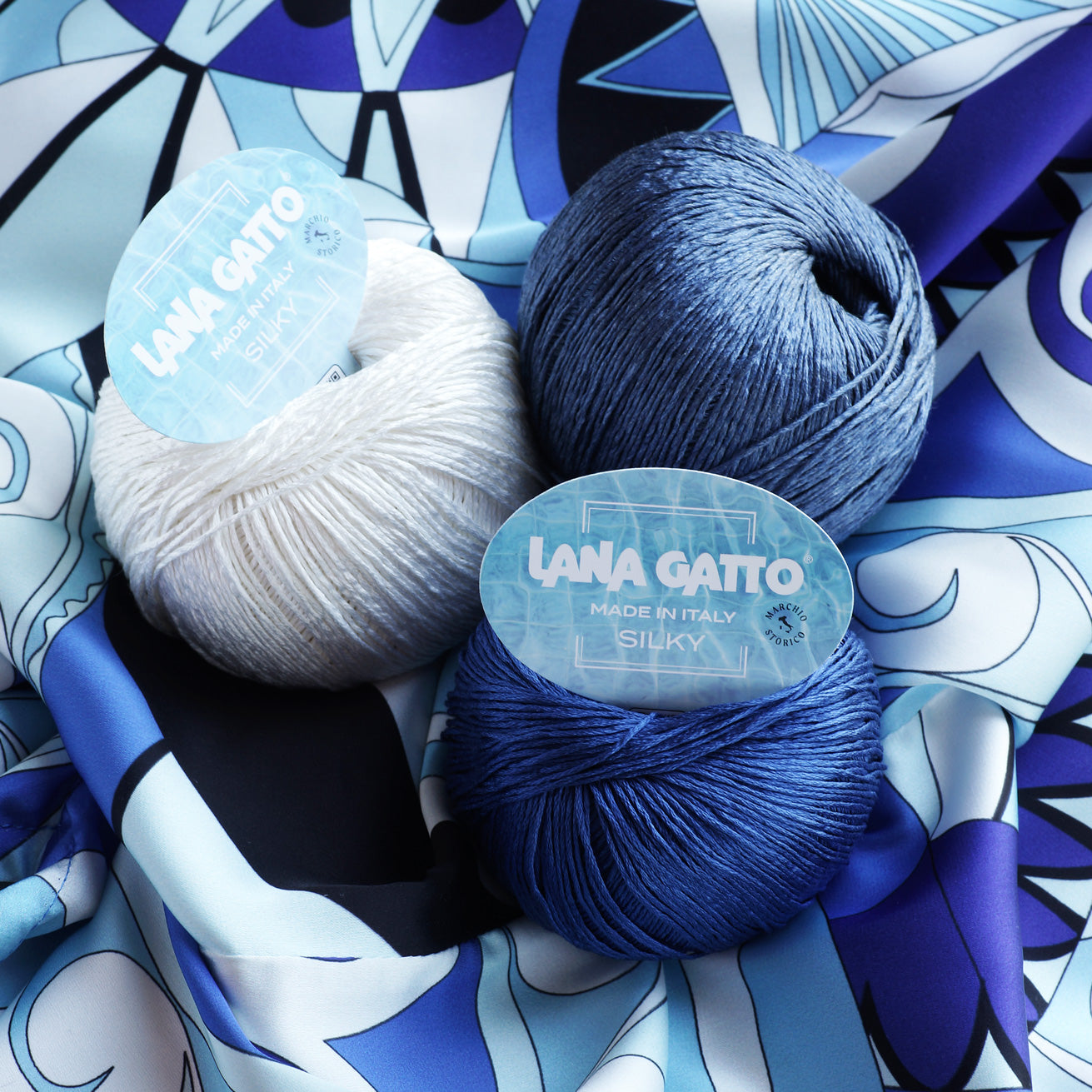 Lana Gatto Silky yarn balls in cream, denim blue, and royal blue on a printed silk fabric