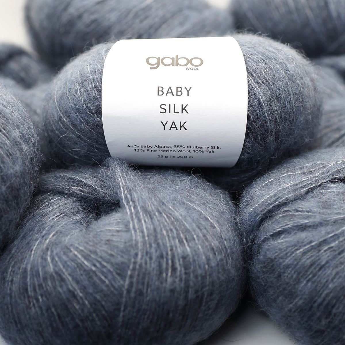 A close-up of a skein of Gabo Wool, Baby Silk Yak yarn in the color 9917 Gray Blue, featuring a white paper label with black text, set against a background of similar gray-blue yarn skeins